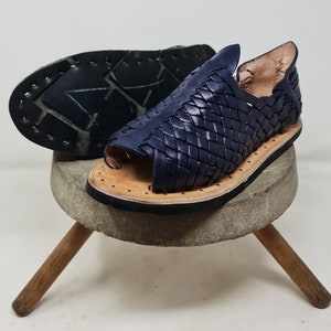 Pu&ograve; includere: Un paio di sandali in pelle blu scuro con un design intrecciato. I sandali hanno la punta chiusa e il tallone aperto. I sandali sono su un supporto in legno.
