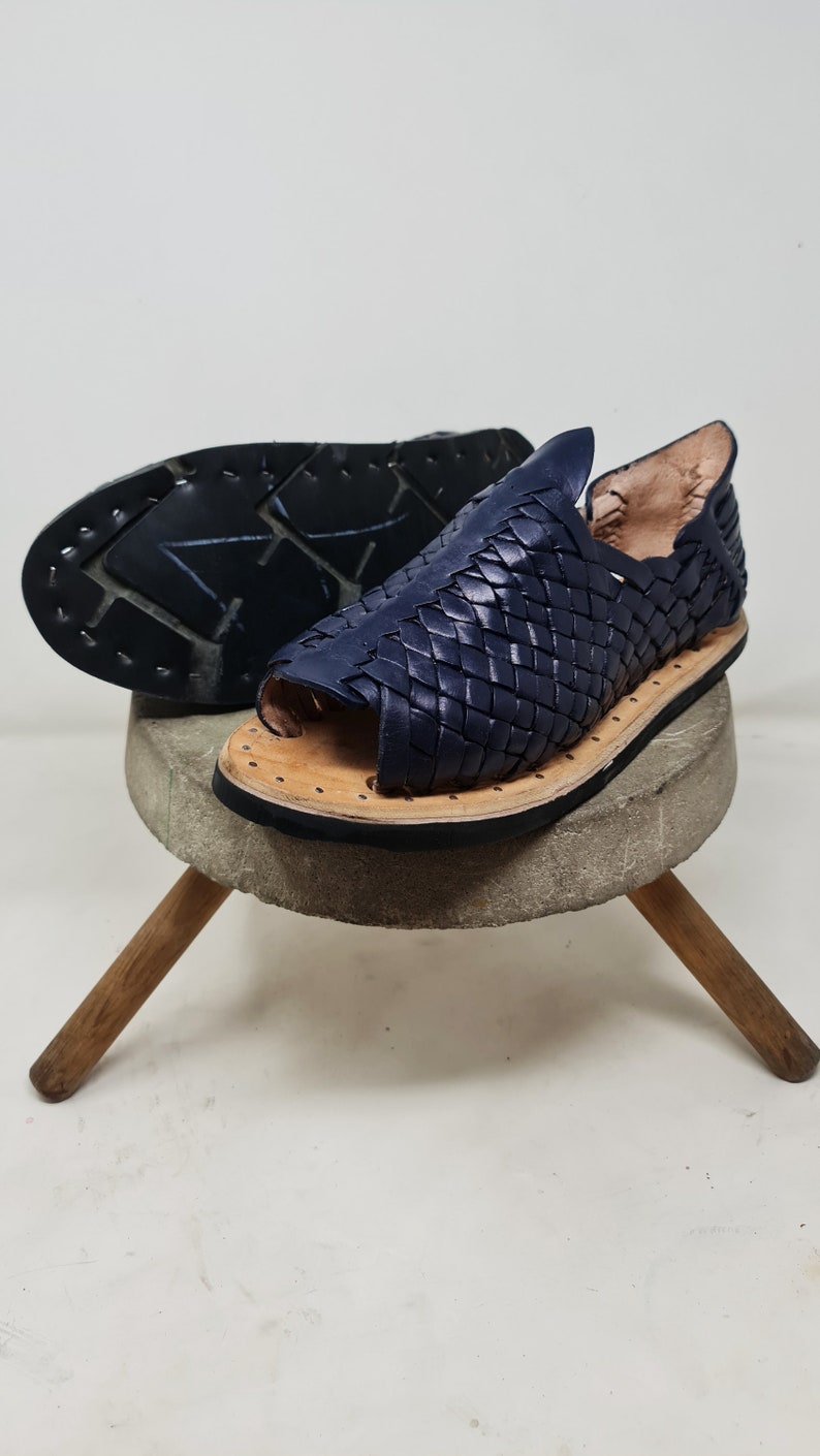 Pu&ograve; includere: Un paio di sandali in pelle blu scuro con un design intrecciato. I sandali hanno la punta chiusa e il tallone aperto. I sandali sono su un supporto in legno.