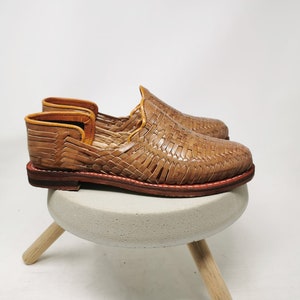 May include: A pair of woven leather sandals in a light brown color. The sandals have a simple design and are made of a soft, supple leather. They are perfect for casual wear and would be comfortable for everyday use.
