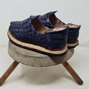 Pu&ograve; includere: Un paio di sandali in pelle blu navy con un design intrecciato. I sandali sono su un piedistallo di cemento con gambe di legno.