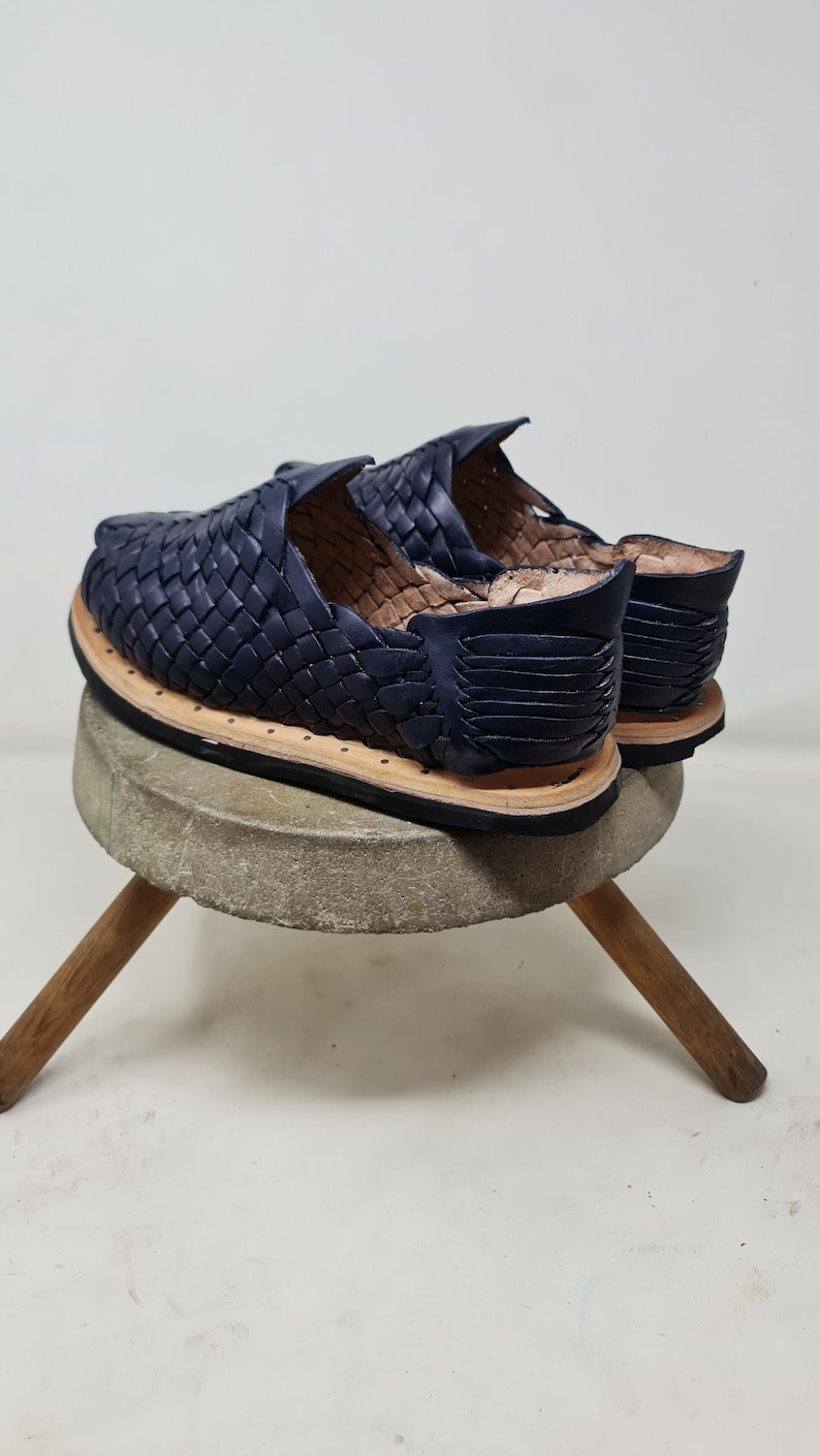 Pu&ograve; includere: Un paio di sandali in pelle blu navy con un design intrecciato. I sandali sono su un piedistallo di cemento con gambe di legno.