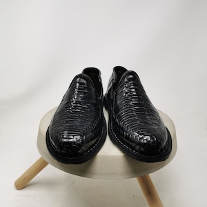 May include: A pair of black leather slip-on shoes with a woven texture. The shoes are displayed on a small, round, concrete-colored stool with wooden legs, set against a white background.