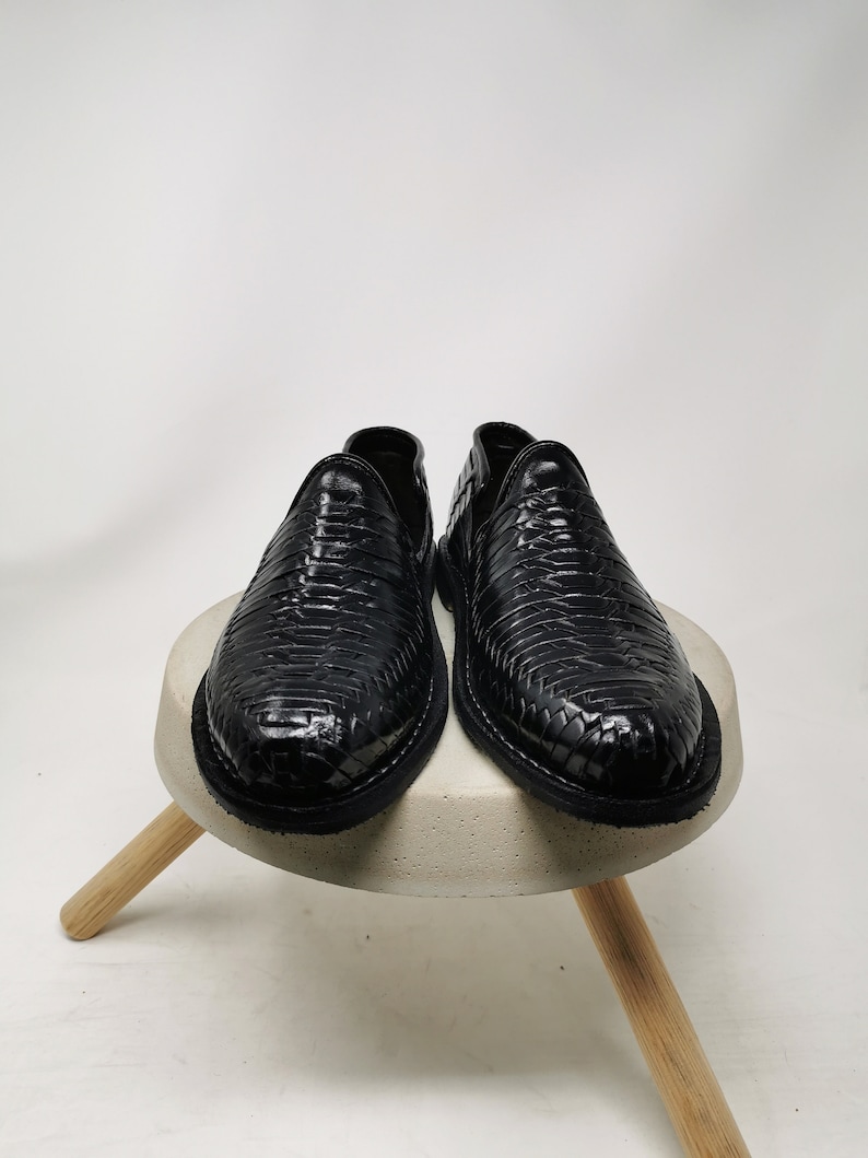 May include: A pair of black leather slip-on shoes with a woven texture. The shoes are displayed on a small, round, concrete-colored stool with wooden legs, set against a white background.