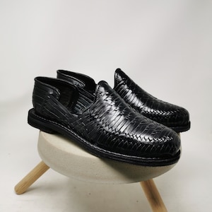 May include: A pair of black leather woven shoes. The shoes are displayed on a small, round, concrete-colored stool with three wooden legs. The shoes have a slip-on design with a textured, woven pattern.