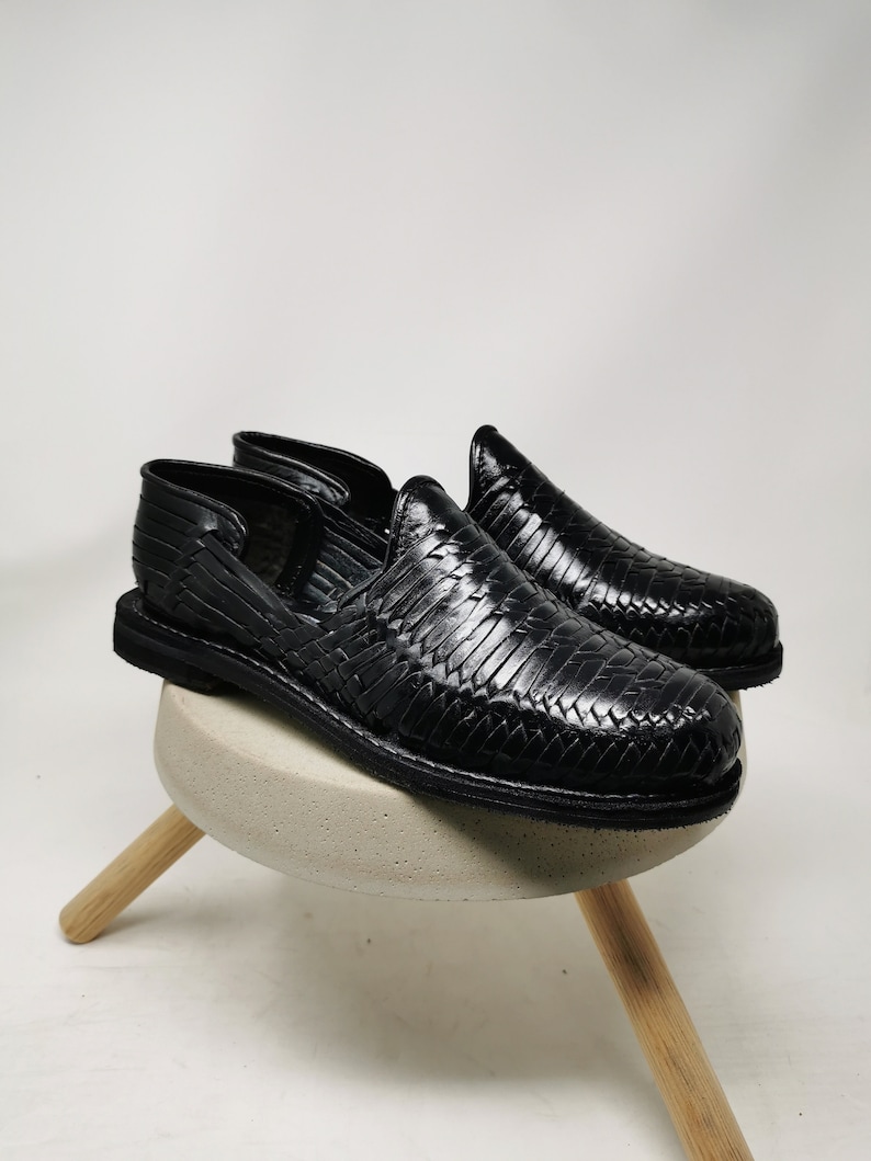 May include: A pair of black leather woven shoes. The shoes are displayed on a small, round, concrete-colored stool with three wooden legs. The shoes have a slip-on design with a textured, woven pattern.