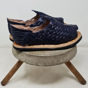Pu&ograve; includere: Un paio di sandali in pelle blu navy con un design intrecciato. I sandali sono su uno sgabello di legno con un piano in pietra grigia.