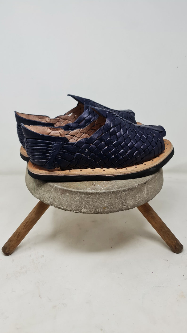 Pu&ograve; includere: Un paio di sandali in pelle blu navy con un design intrecciato. I sandali sono su uno sgabello di legno con un piano in pietra grigia.