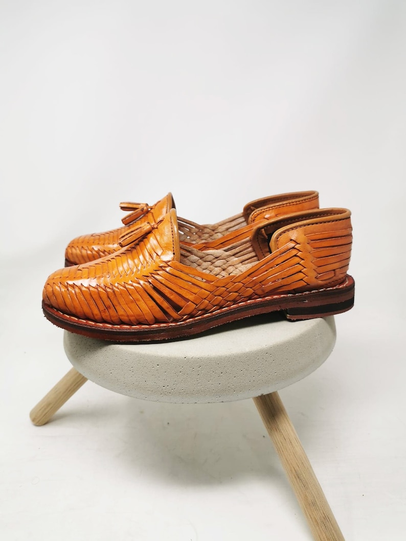 Puede incluir: Un par de sandalias de cuero color canela con un dise&ntilde;o tejido y un detalle de borla. Las sandalias est&aacute;n sobre un peque&ntilde;o taburete de madera.