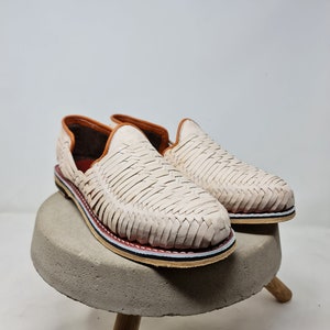 Mocasines de cuero crema con diseño trenzado y suela de esparto multicolor, sobre base circular de concreto, transmitiendo un estilo elegante y artesanal.