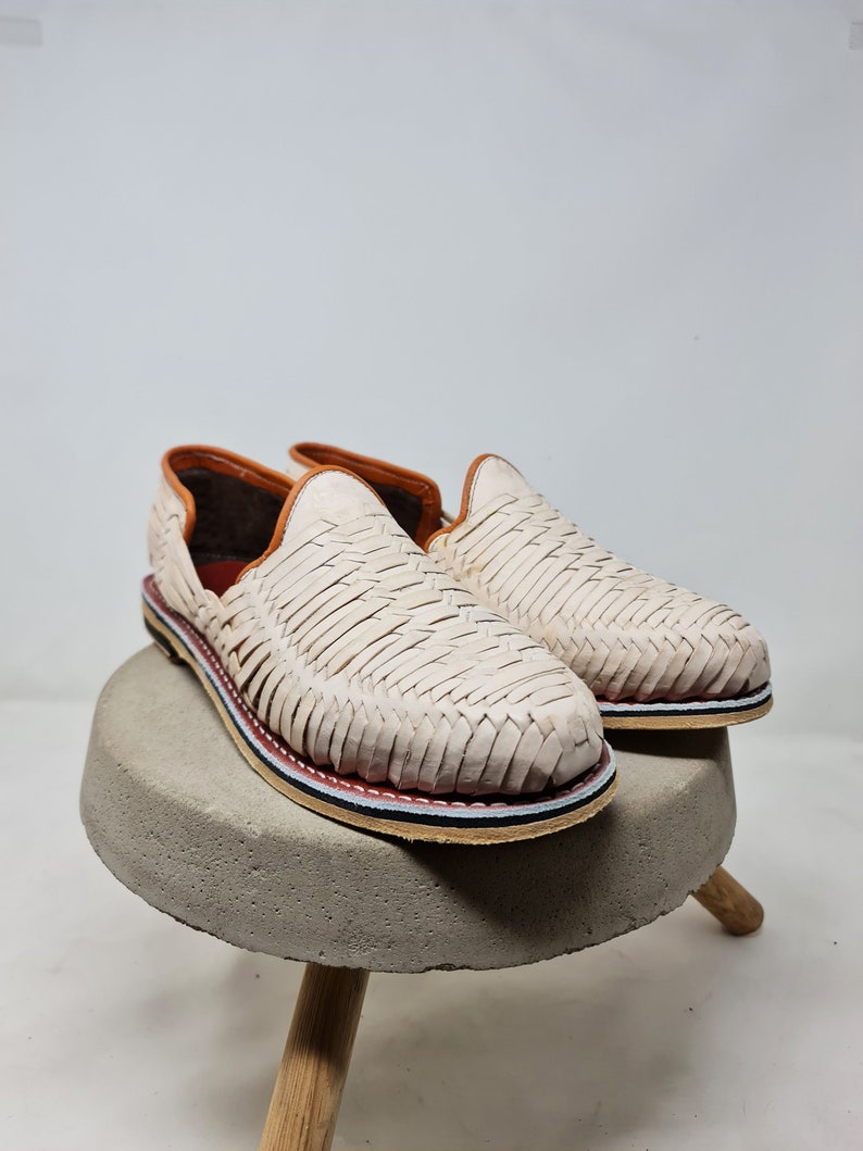 Mocasines de cuero crema con diseño trenzado y suela de esparto multicolor, sobre base circular de concreto, transmitiendo un estilo elegante y artesanal.