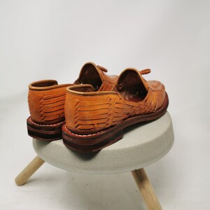 Puede incluir: Un par de sandalias de cuero marr&oacute;n con correas tejidas. Las sandalias est&aacute;n sobre un peque&ntilde;o taburete de hormig&oacute;n con patas de madera.