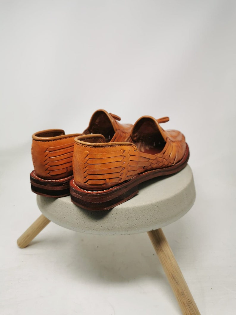 Puede incluir: Un par de sandalias de cuero marr&oacute;n con correas tejidas. Las sandalias est&aacute;n sobre un peque&ntilde;o taburete de hormig&oacute;n con patas de madera.