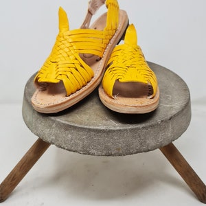 May include: A pair of yellow leather sandals with a woven design. The sandals are on a gray stone pedestal with wooden legs.