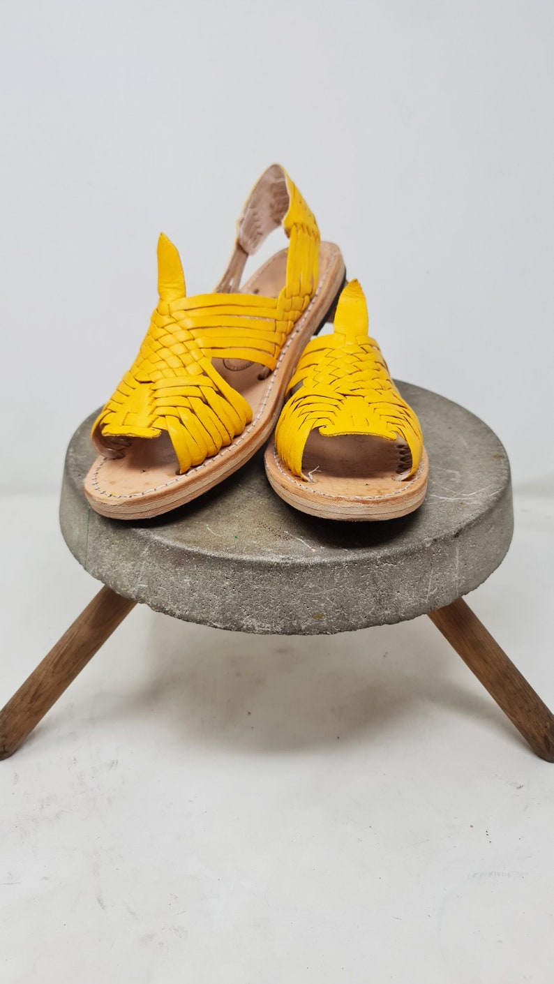 May include: A pair of yellow leather sandals with a woven design. The sandals are on a gray stone pedestal with wooden legs.
