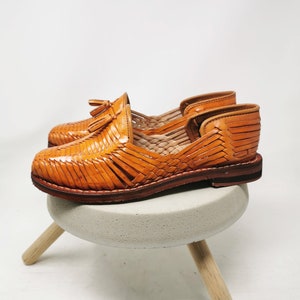 Puede incluir: Un par de sandalias de cuero color canela con un dise&ntilde;o tejido y un detalle de borla. Las sandalias est&aacute;n sobre un peque&ntilde;o taburete de madera.