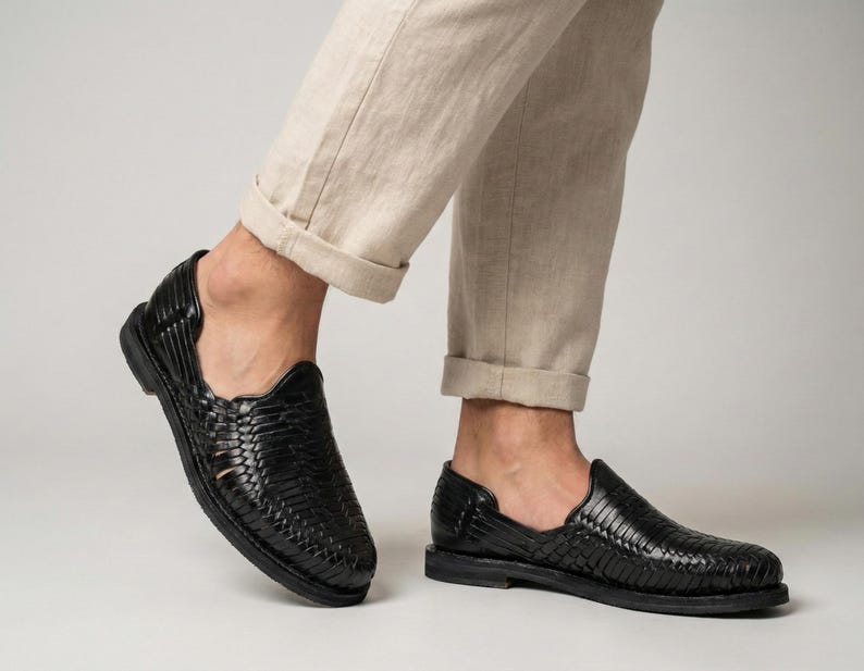 May include: Black leather woven slip-on shoes. The shoes have a rounded toe and a low heel. The woven design covers the entire shoe, with a small opening on the side. The shoes are worn with beige pants.