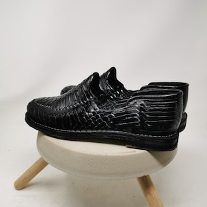 May include: A pair of black leather woven shoes. The shoes are displayed on a small, round, light gray stool with three wooden legs. The shoes have a textured, woven design and a low profile sole.