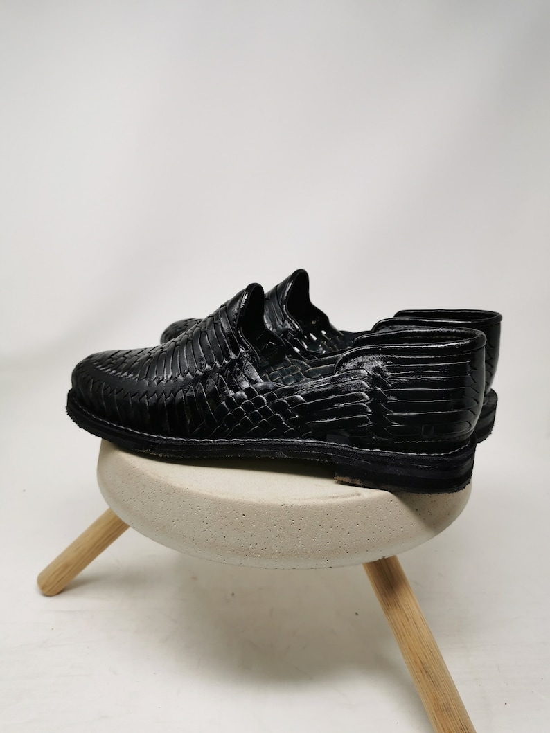 May include: A pair of black leather woven shoes. The shoes are displayed on a small, round, light gray stool with three wooden legs. The shoes have a textured, woven design and a low profile sole.