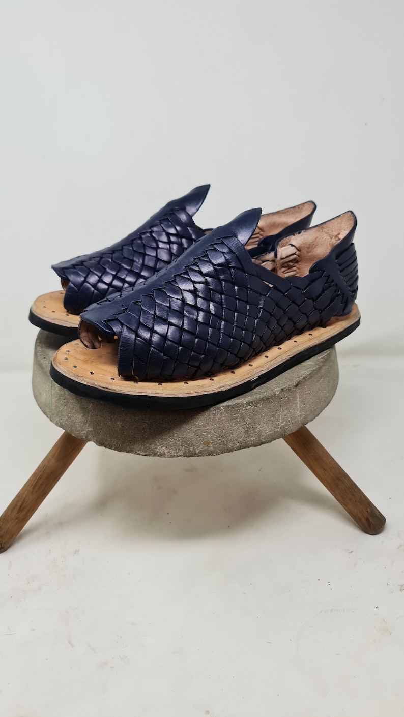 Pu&ograve; includere: Un paio di sandali in pelle blu navy con un design intrecciato. I sandali sono su uno sgabello di legno.