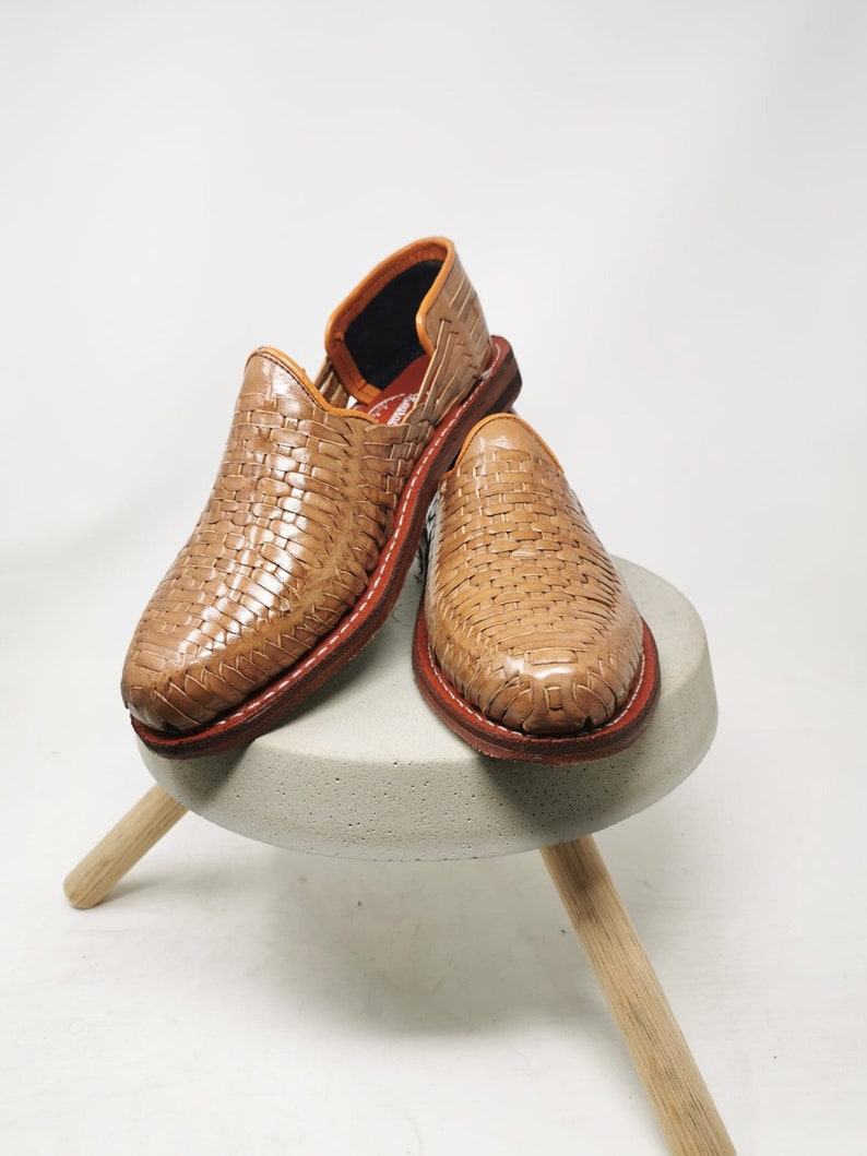 May include: A pair of woven leather shoes in a light brown hue, with a red stitched sole. The shoes are displayed on a small, round, gray concrete stool with wooden legs. The shoes have a classic, slip-on design.
