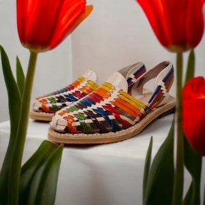 May include: A pair of colorful woven leather sandals with a rainbow pattern. The sandals have a strap that goes around the ankle and a toe strap. The sandals are on a white surface.