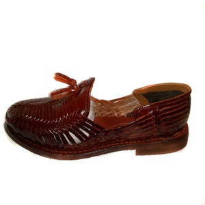 May include: A brown leather sandal with a woven design. The sandal features a small tassel detail and a low heel. The shoe is designed with open sides and a closed toe. The sandal is approximately 10 inches long.