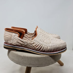 Mocasines artesanales de cuero trenzado color crema, perfilados con suela multicolor y reposando sobre pedestal de hormigón, capturando la fusión de tradición y diseño moderno.