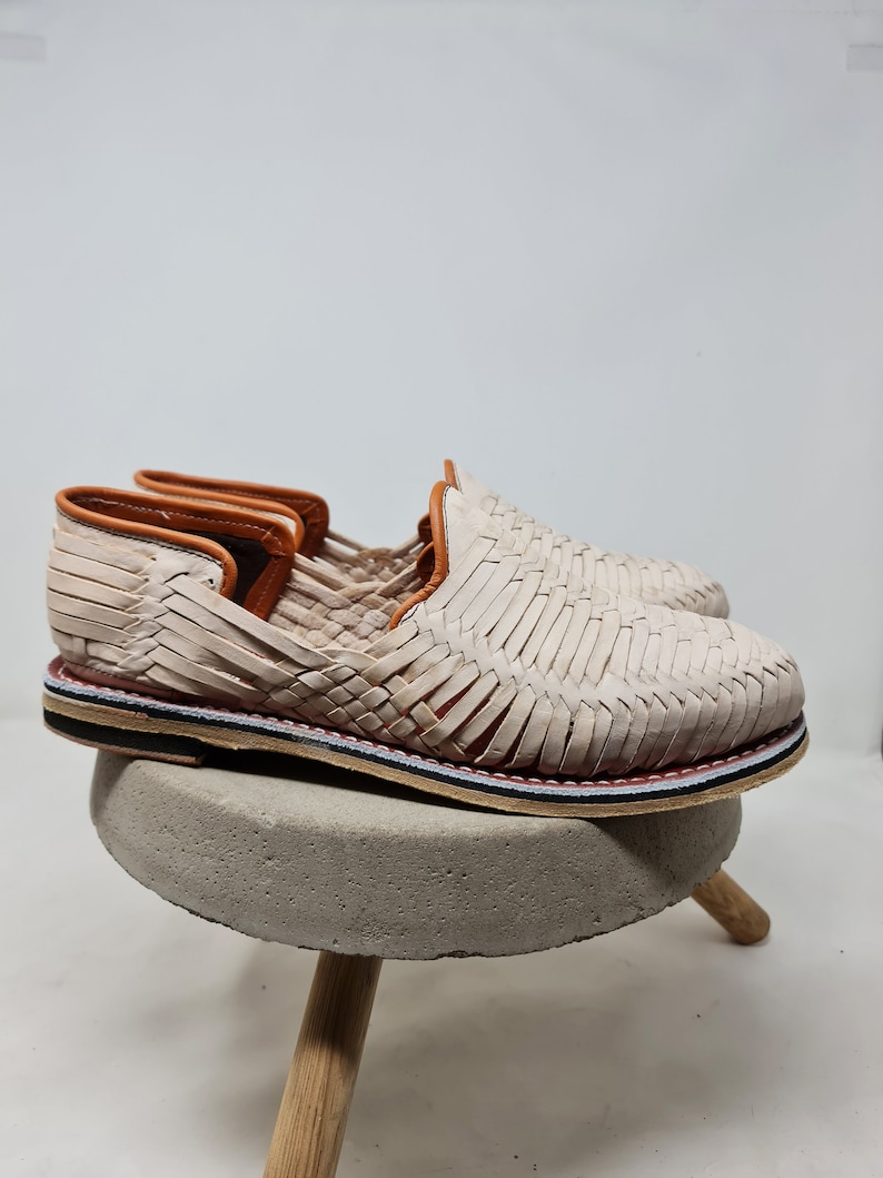 Mocasines artesanales de cuero trenzado color crema, perfilados con suela multicolor y reposando sobre pedestal de hormigón, capturando la fusión de tradición y diseño moderno.