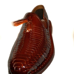 May include: A brown leather huarache shoe with a woven design and a small tassel. The shoe has a glossy finish and a rounded toe. The shoe is a slip-on style.