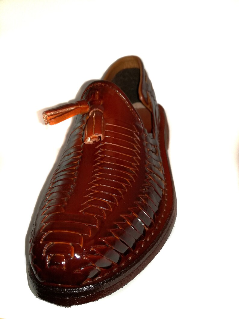 May include: A brown leather huarache shoe with a woven design and a small tassel. The shoe has a glossy finish and a rounded toe. The shoe is a slip-on style.