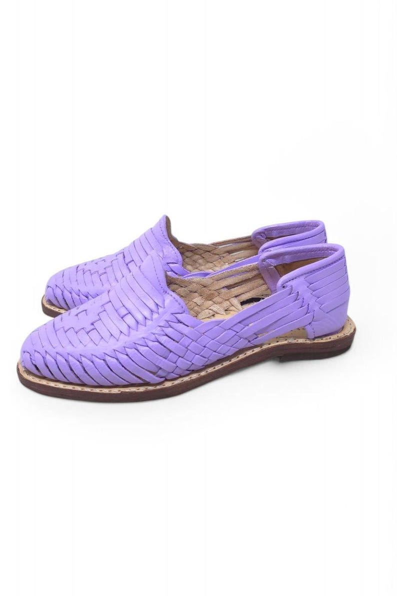 May include: A pair of lavender huarache sandals. These woven leather shoes have a closed toe and heel with an open side design. The soles are brown with a light-colored stitching.