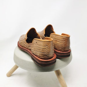 May include: A pair of tan leather huaraches, featuring woven uppers and brown soles with white stitching. The shoes are displayed on a small, light gray stool with wooden legs, against a white background.