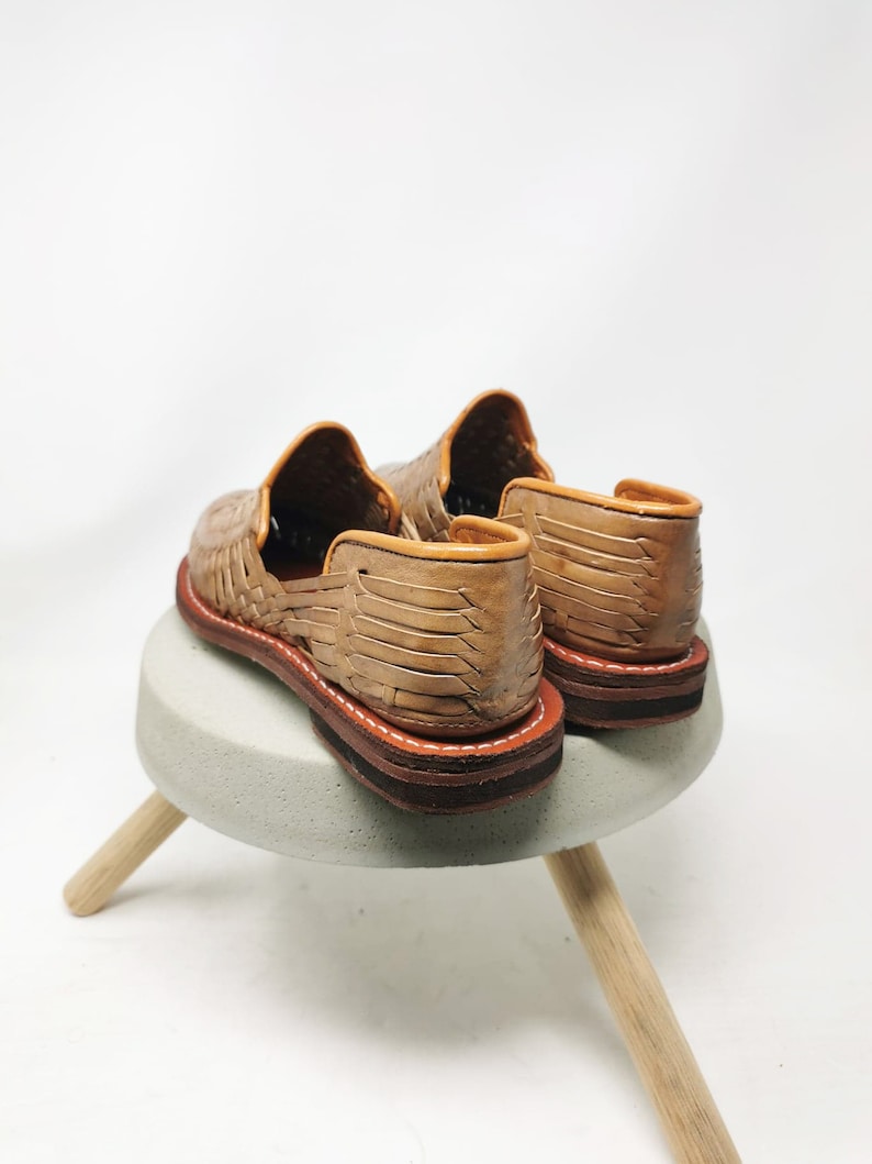 May include: A pair of tan leather huaraches, featuring woven uppers and brown soles with white stitching. The shoes are displayed on a small, light gray stool with wooden legs, against a white background.