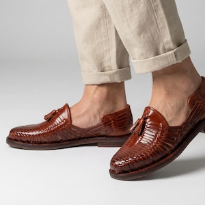 Könnte beinhalten: Braune, geflochtene Lederschuhe mit Quasten-Detail. Die Schuhe haben ein niedriges Profil und eine flache Sohle. Das Bild zeigt die Schuhe aus einem niedrigen Winkel, wobei die Beine des Trägers ab den Knien abwärts sichtbar sind.