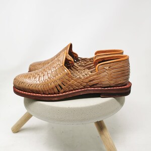 May include: A pair of woven leather shoes in a light brown hue. The shoes feature a woven design on the upper and a brown sole with red stitching. The shoes are displayed on a small, round, gray stool with wooden legs.