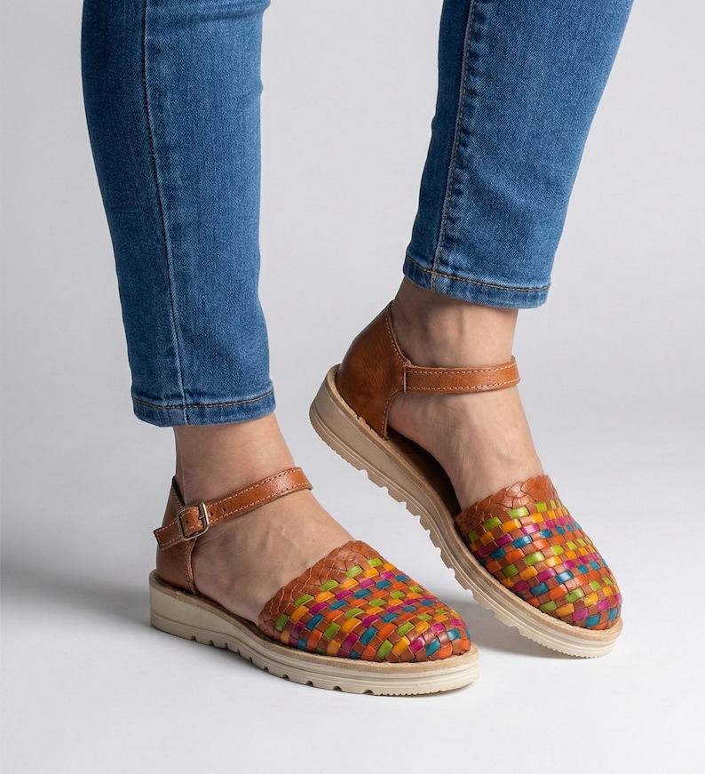 Puede incluir: Sandalias de cuero marr&oacute;n con puntera tejida multicolor. Las sandalias tienen suela beige y correa de tobillo con hebilla. Los zapatos se llevan con vaqueros azules.
