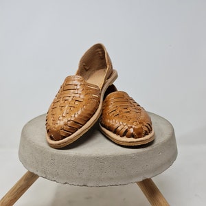 Puede incluir: Un par de sandalias de cuero tejido en un tono marr&oacute;n claro. Las sandalias se exhiben sobre un peque&ntilde;o taburete redondo de hormig&oacute;n gris con patas de madera. Las sandalias tienen suela plana y dise&ntilde;o abierto.