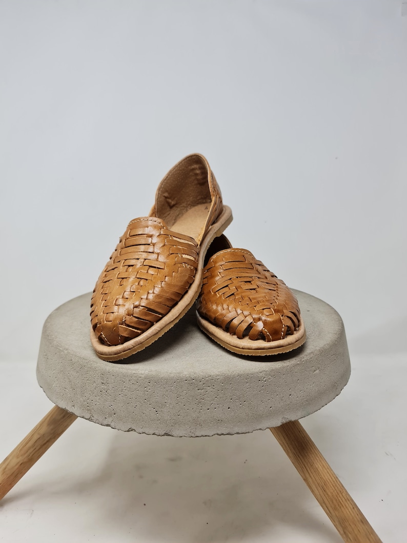 Puede incluir: Un par de sandalias de cuero tejido en un tono marr&oacute;n claro. Las sandalias se exhiben sobre un peque&ntilde;o taburete redondo de hormig&oacute;n gris con patas de madera. Las sandalias tienen suela plana y dise&ntilde;o abierto.