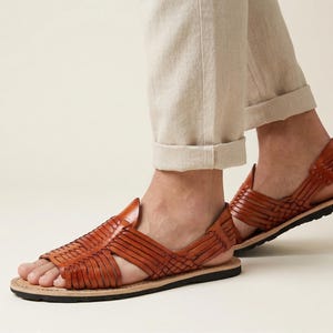 May include: Brown leather sandals with a woven design. The sandals have a flat sole and a strap around the ankle. The sandals are worn with beige trousers. The sandals are a size 9.