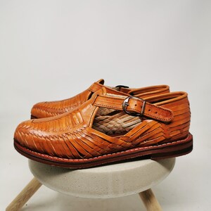 May include: A pair of handcrafted, open-toe leather sandals in a warm, tan color. The sandals feature a woven design across the top and a buckle closure. The soles are a darker brown, and the shoes are displayed on a small, light-colored stool.