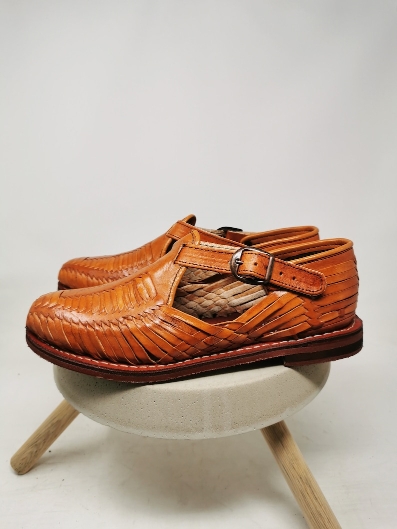 May include: A pair of handcrafted, open-toe leather sandals in a warm, tan color. The sandals feature a woven design across the top and a buckle closure. The soles are a darker brown, and the shoes are displayed on a small, light-colored stool.