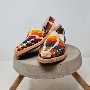 May include: A pair of colorful leather sandals with a woven design. The sandals are on a concrete pedestal with wooden legs.
