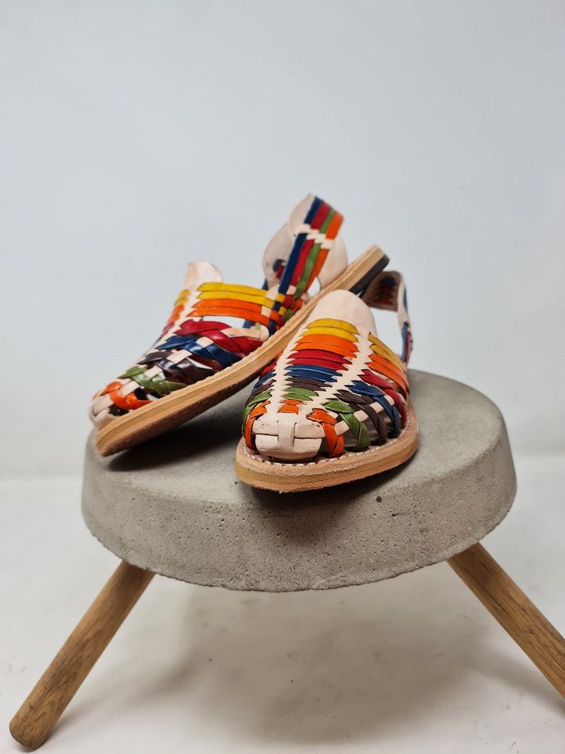 May include: A pair of colorful leather sandals with a woven design. The sandals are on a concrete pedestal with wooden legs.