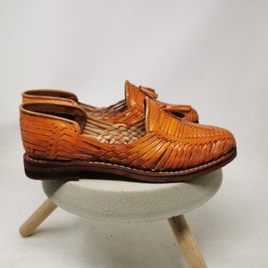 Puede incluir: Un par de sandalias de cuero marr&oacute;n con un dise&ntilde;o tejido. Las sandalias tienen un detalle de borla y una suela de cuero.