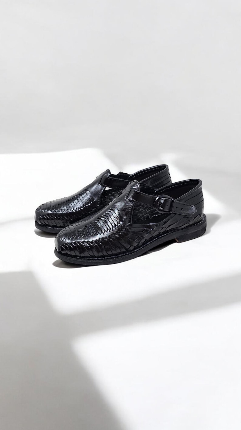 May include: A pair of black leather huarache sandals. The sandals feature a woven design across the top and a buckle closure on the side. The shoes are set against a white background.