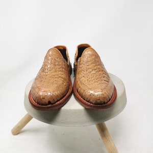 May include: A pair of light brown woven leather shoes. The shoes are displayed on a small, round, gray concrete stool with three wooden legs. The shoes have a woven pattern and a brown leather sole.