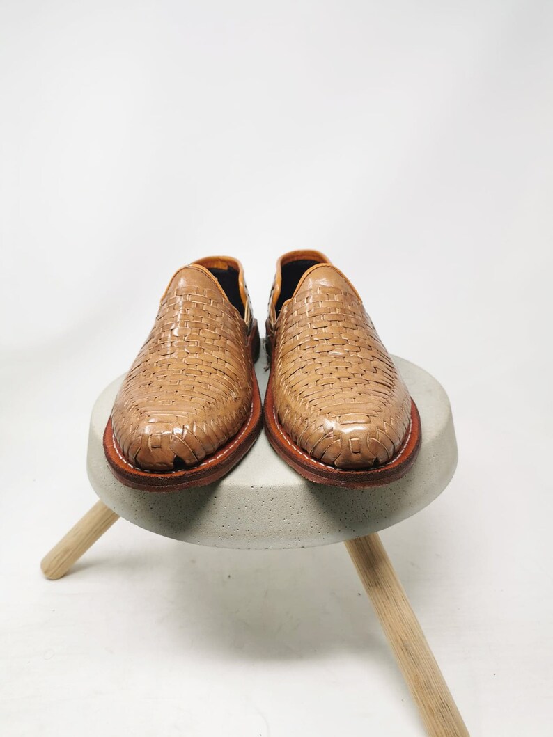 May include: A pair of light brown woven leather shoes. The shoes are displayed on a small, round, gray concrete stool with three wooden legs. The shoes have a woven pattern and a brown leather sole.