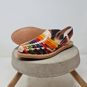 May include: A pair of colorful leather sandals with a woven design. The sandals have a white leather base and are decorated with stripes of red, orange, yellow, green, blue, and brown leather. The sandals are sitting on a concrete pedestal with wooden legs.