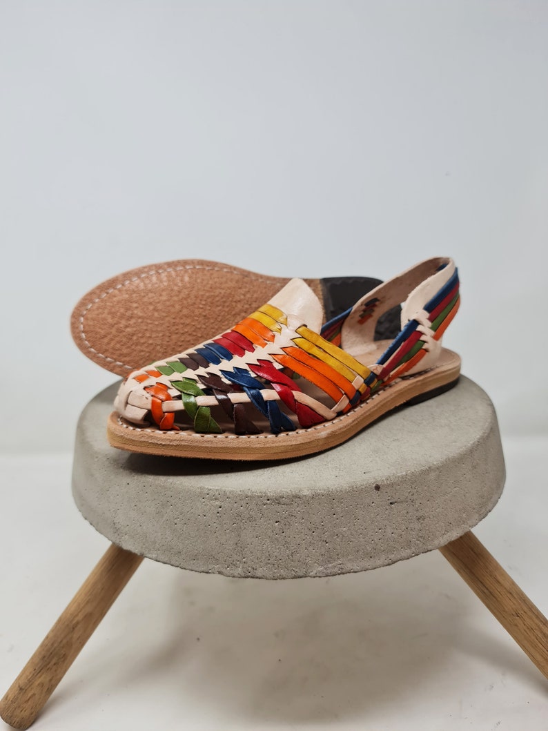 May include: A pair of colorful leather sandals with a woven design. The sandals have a white leather base and are decorated with stripes of red, orange, yellow, green, blue, and brown leather. The sandals are sitting on a concrete pedestal with wooden legs.