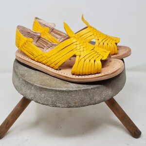 May include: A pair of yellow leather sandals with a woven design. The sandals are sitting on a wooden stool.